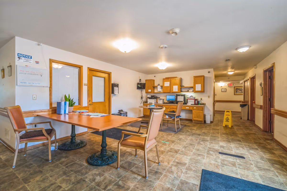 Interior view of a senior living facility office area with a large wooden table surrounded by chairs, a desk with computers and office supplies against the back wall, and a hallway leading to other rooms. The floor is tiled, and the walls are light-colored with wooden trim. There are ceiling lights and a caution wet floor sign in the hallway.