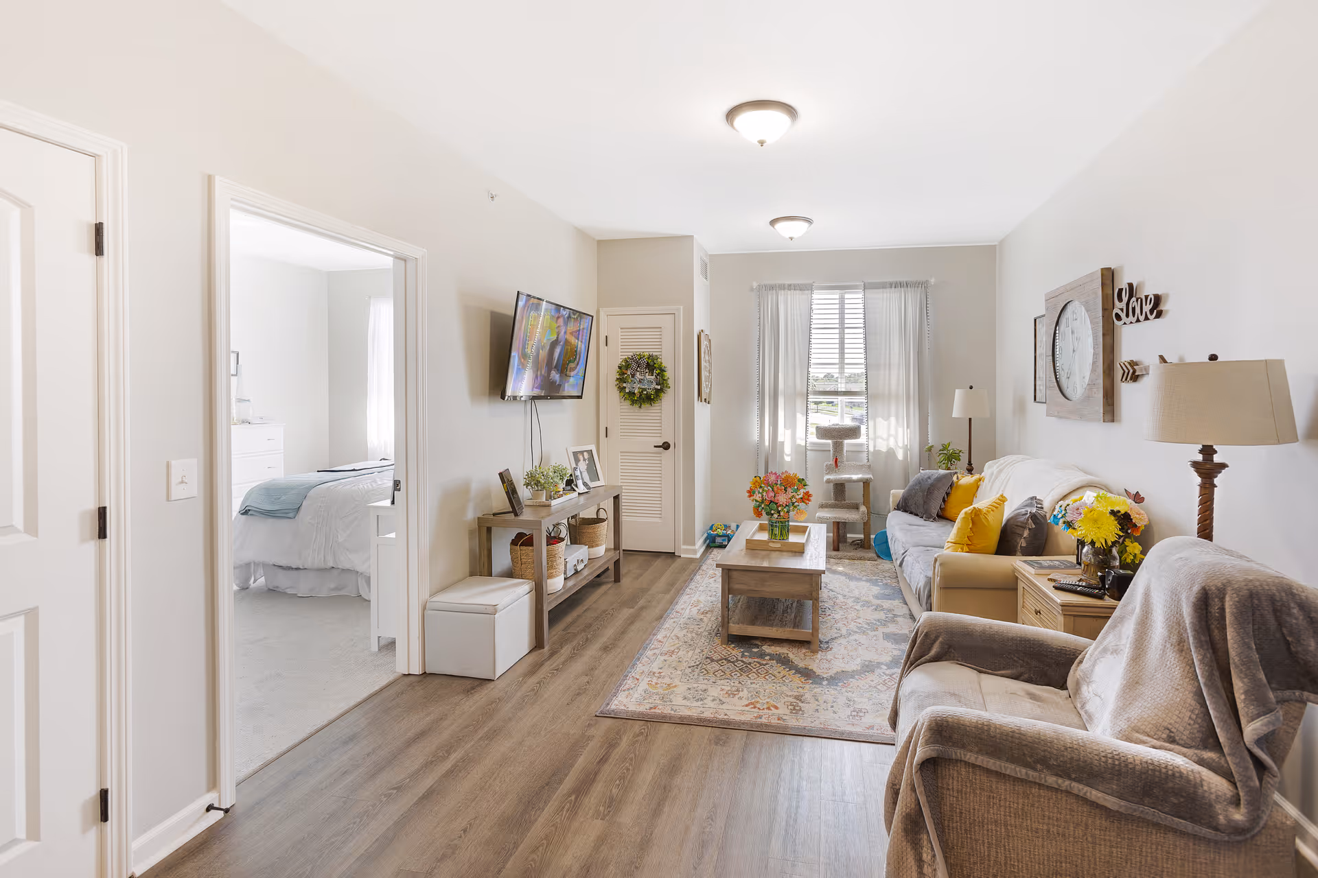 A bright and cozy living room with a beige sofa adorned with yellow and gray pillows, a wooden coffee table with a vase of flowers, a gray armchair with a throw blanket, a floor lamp, and a wall clock with the word 'Love' on the wall. A TV is mounted on the wall opposite the sofa, and a doorway leads to a bedroom with a neatly made bed. The room has wood flooring and a window with sheer curtains allowing natural light to fill the space.