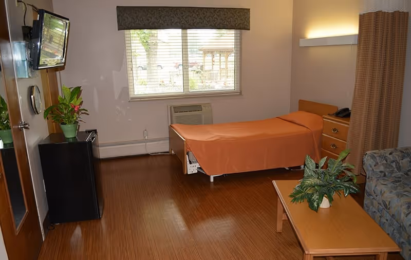 A nursing center room with a single bed covered in an orange bedspread, a wooden nightstand with a telephone, a window with blinds and a valance, a wall-mounted TV, a small black refrigerator with a potted plant on top, a wooden coffee table with a green potted plant, and a patterned armchair. The room has wooden flooring and neutral-colored walls.