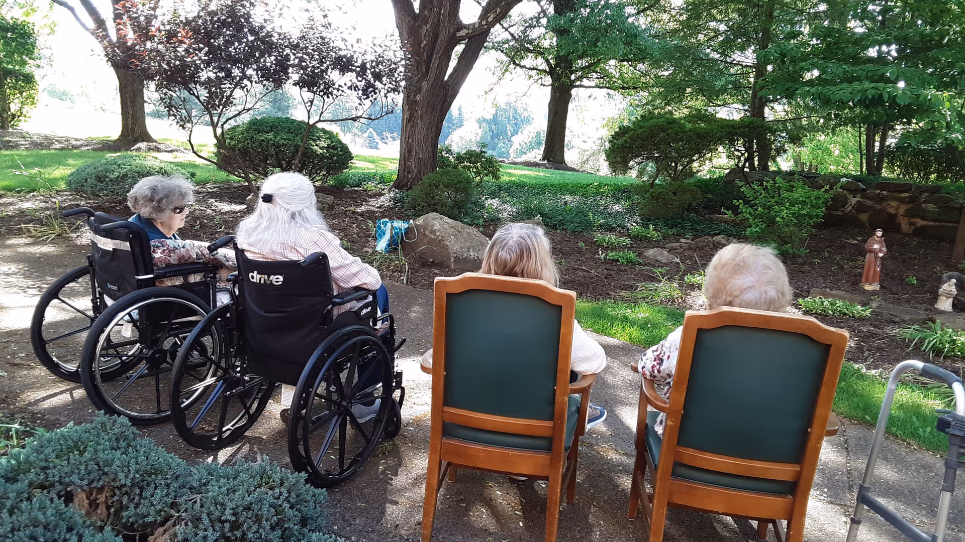 Four elderly individuals sitting outdoors in a garden area; two are in wheelchairs and two are seated in wooden chairs with green cushions, surrounded by trees, bushes, and a walking aid nearby.