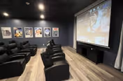 A small theater room with multiple black recliner chairs arranged in rows facing a large projection screen on a dark blue wall. The floor is wooden, and there are framed movie posters on the left wall. The room has a cozy and modern ambiance with ceiling lights.