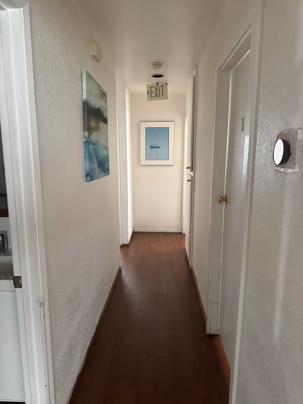A narrow hallway with wooden flooring and white walls. There are doors on both sides of the hallway, a framed picture of a boat at the end, and an exit sign hanging from the ceiling. A piece of abstract art is hung on the left wall.