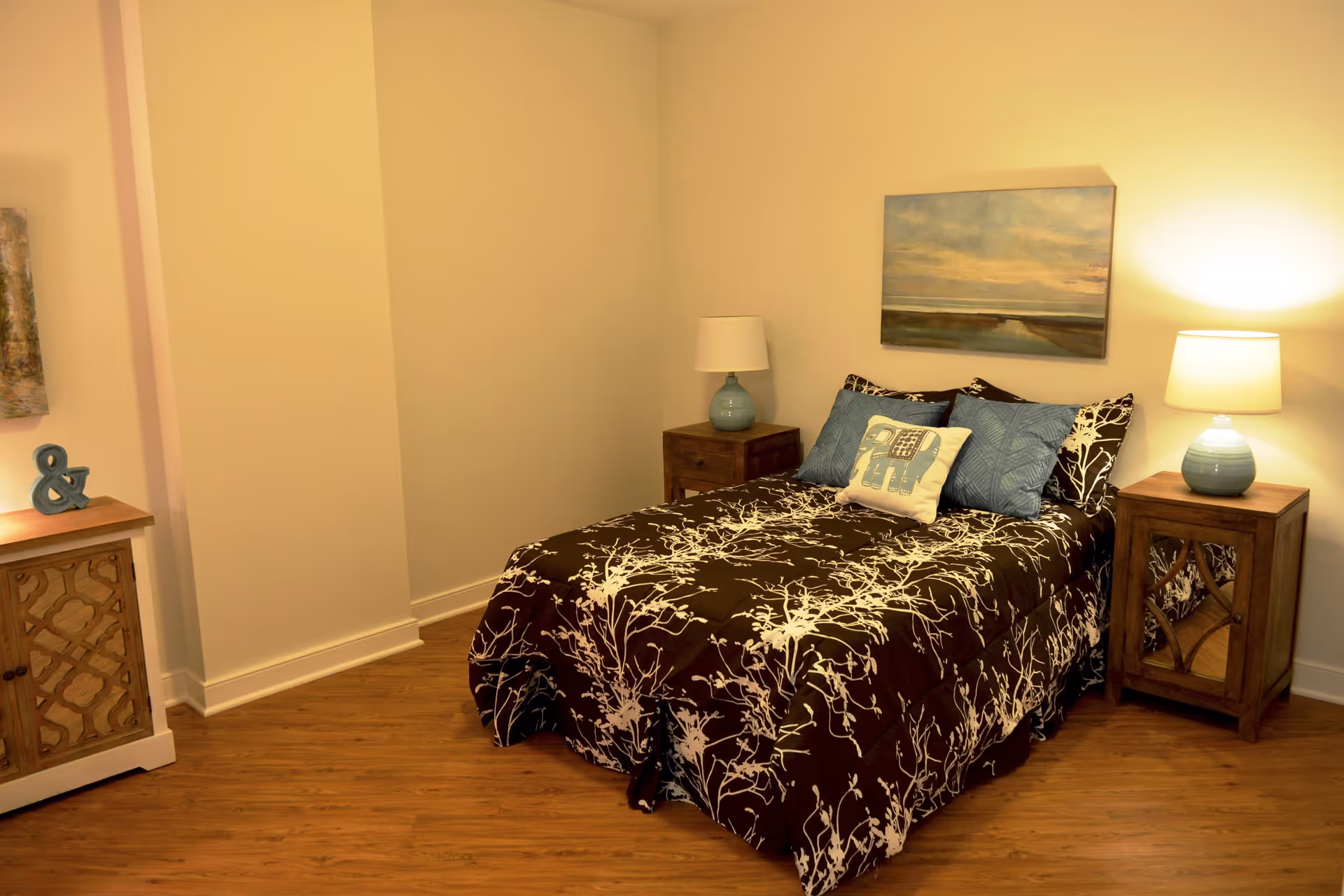 A cozy bedroom with a bed covered in a brown and white patterned comforter, two blue pillows, and a decorative pillow with an elephant design. There are two wooden nightstands on either side of the bed, each with a blue lamp. A landscape painting hangs on the wall above the bed. The room has wooden flooring and light-colored walls.