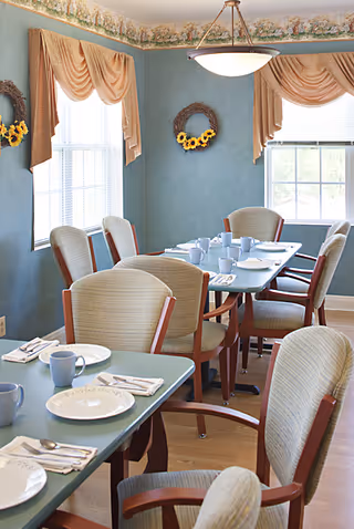 Small dining room set for a meal with tables, cups and plates, upholstered chairs, blue-green walls, windows with draped valances and sunflower wreaths.