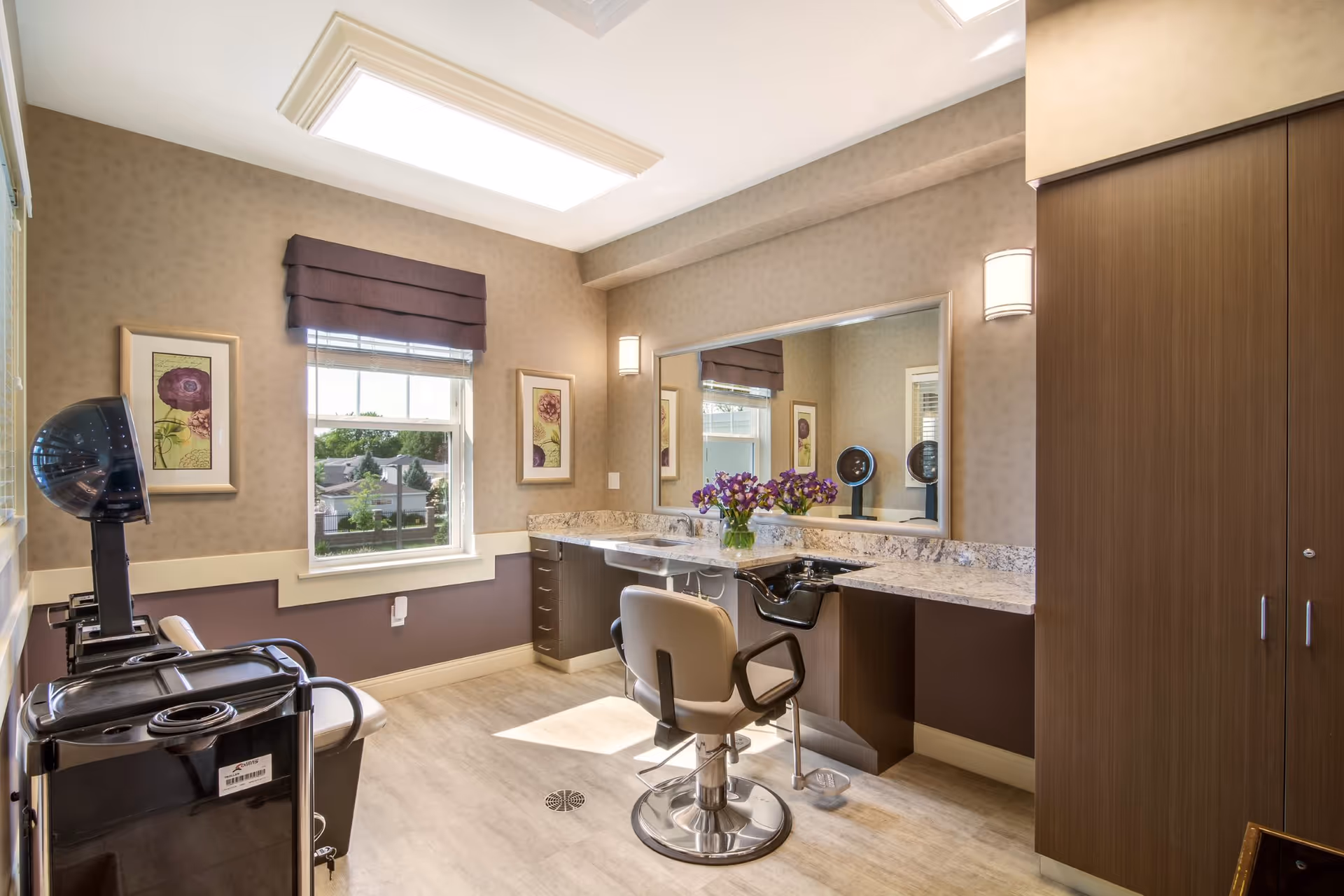 A well-lit salon room with a styling chair in front of a large mirror, a countertop with a vase of purple flowers, two framed floral artworks on the wall, a window with a brown valance, and salon equipment including a hair dryer and a rolling cart.