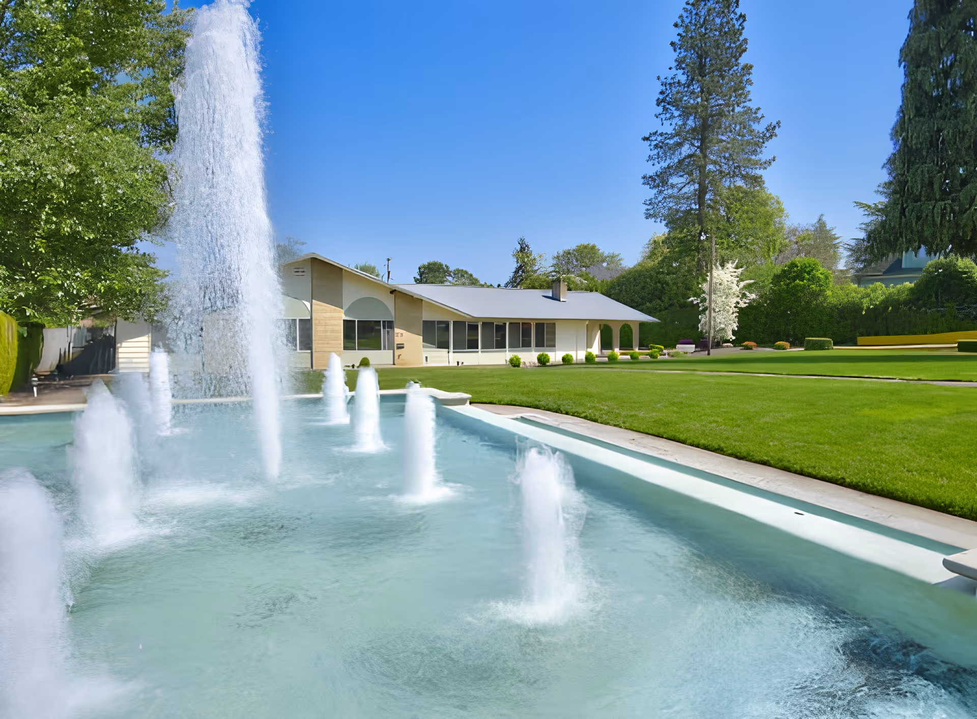 Outdoor view of Jefferson Lodge Memory Care Community featuring a large water fountain with multiple jets spraying water, a well-maintained green lawn, trees, and a single-story building in the background under a clear blue sky.