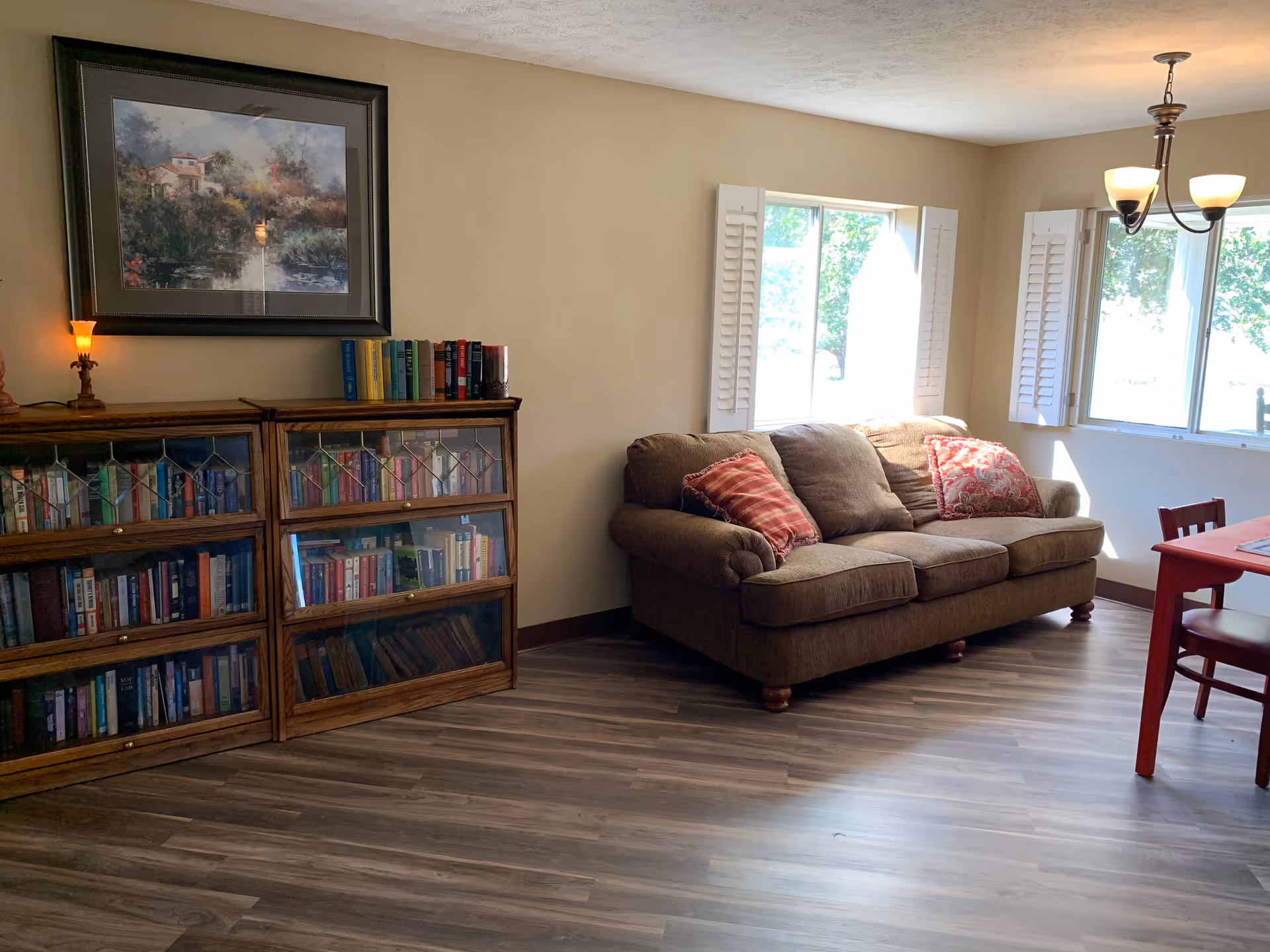 A cozy living room area with a brown sofa adorned with two patterned pillows, a wooden bookshelf filled with books, a framed painting above the bookshelf, and two windows with white shutters letting in natural light. A red table with chairs is partially visible on the right side under a ceiling light fixture.