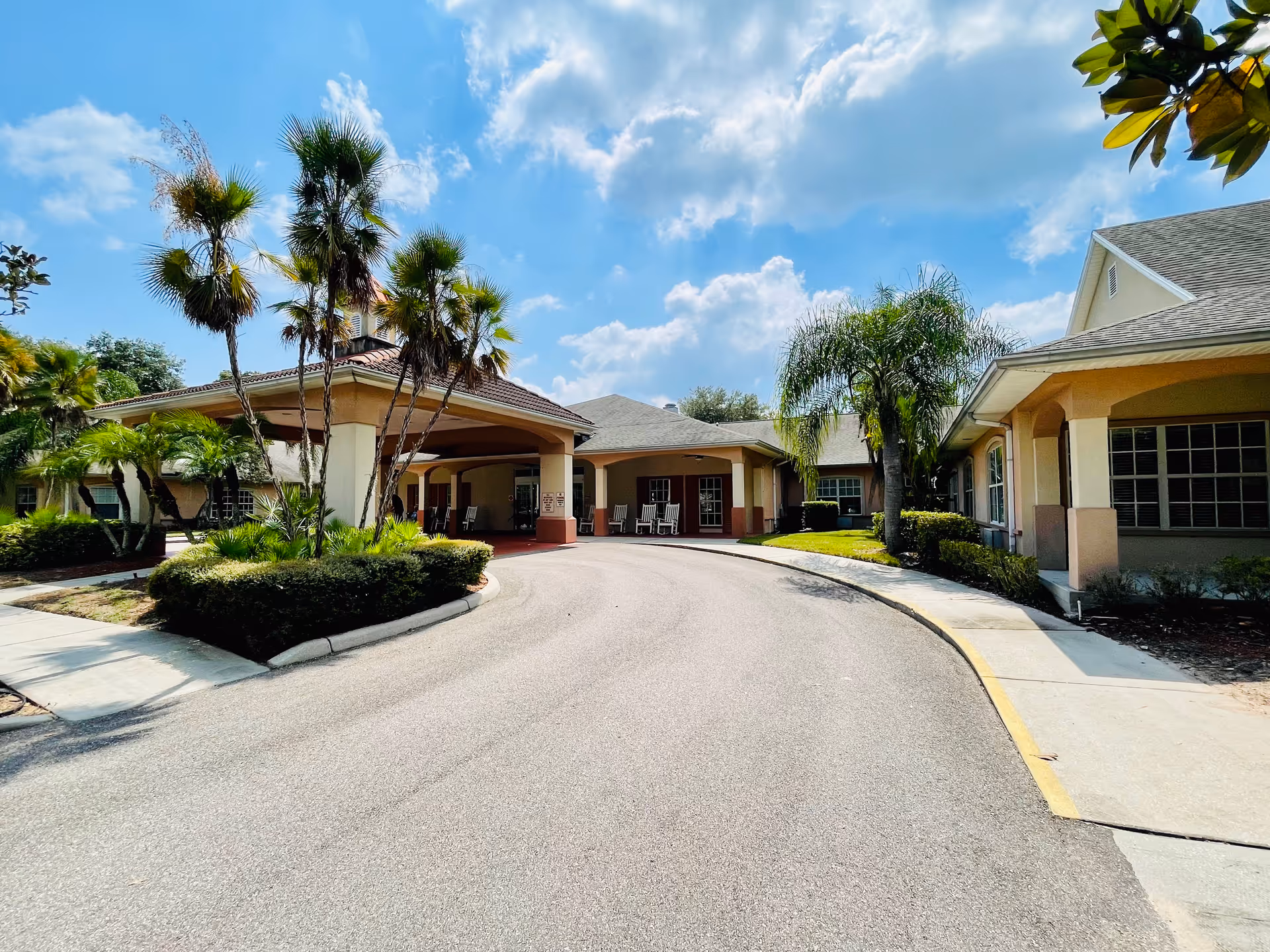 Front exterior view of Hawthorne Estates of Brandon (Independent Living) showing a curved driveway leading to a covered entrance with palm trees and landscaping on either side under a partly cloudy blue sky.