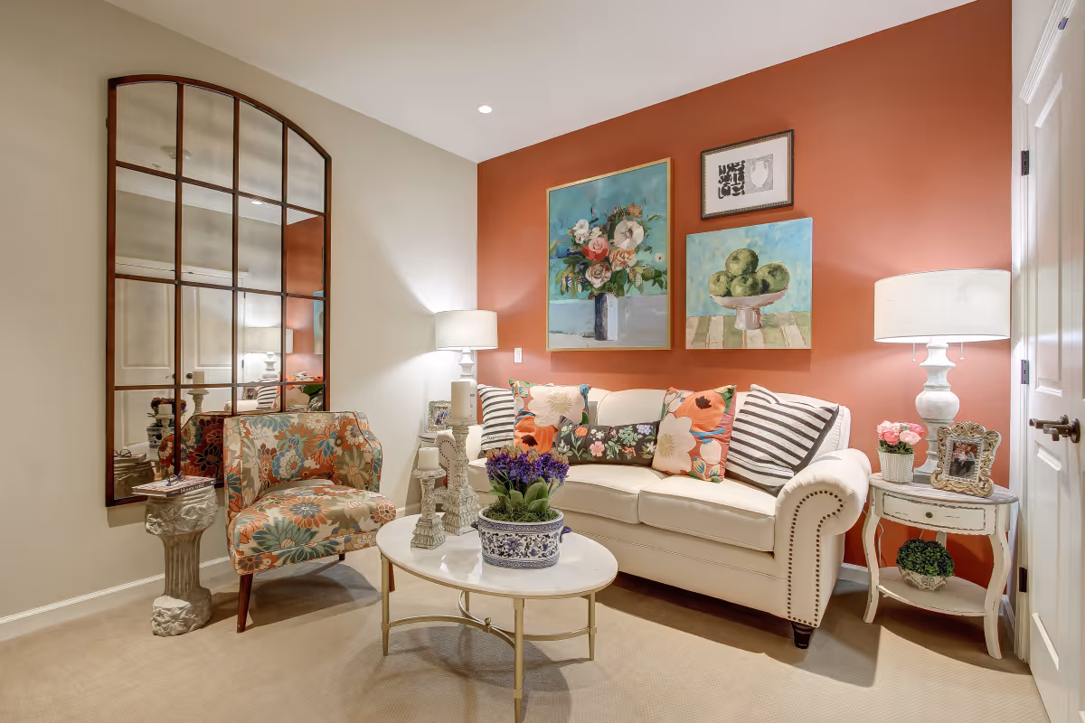 A cozy living room with a white sofa adorned with colorful floral and striped pillows, a floral patterned armchair, a round white coffee table with decorative items and a potted plant, two table lamps on either side, and three paintings on a warm terracotta accent wall. A large windowpane-style mirror is mounted on the adjacent beige wall.