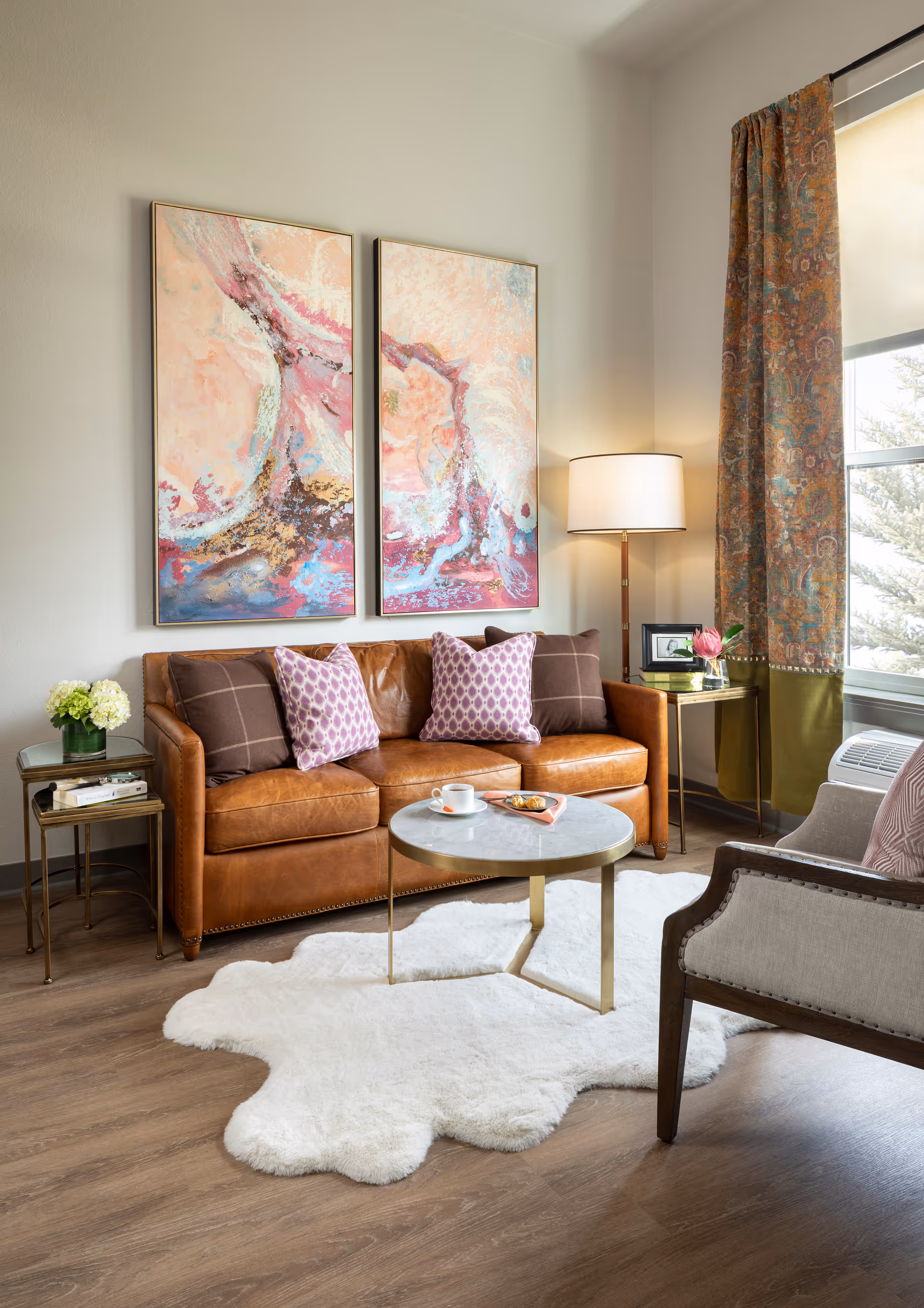 A cozy living room with a brown leather sofa adorned with four decorative pillows, two in purple patterns and two in brown plaid. Above the sofa, there are two large abstract paintings with pink, blue, and beige tones. A round marble coffee table with gold legs sits on a white fluffy rug in front of the sofa, holding a cup and saucer and a small plate with food. To the right, a floor lamp with a white shade stands next to a window with patterned curtains. A beige armchair with a pink pillow is partially visible on the right side. The floor is wooden, and there are two small side tables with books and a flower vase on the left side of the sofa.