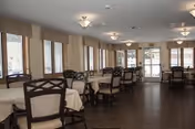 A spacious dining room with multiple tables covered with beige tablecloths and surrounded by wooden chairs with cushioned seats. Large windows with beige valances allow natural light to fill the room. Ceiling lights provide additional illumination, and a glass door is visible at the far end of the room.