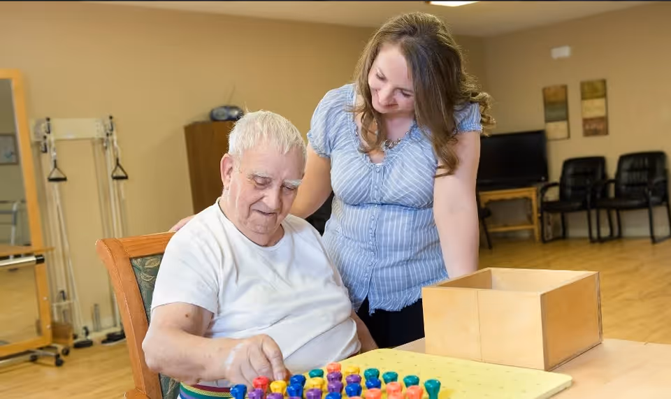 An elderly man sitting at a table playing a colorful pegboard game while a woman stands beside him smiling and offering support in a room with wooden flooring and chairs in the background.