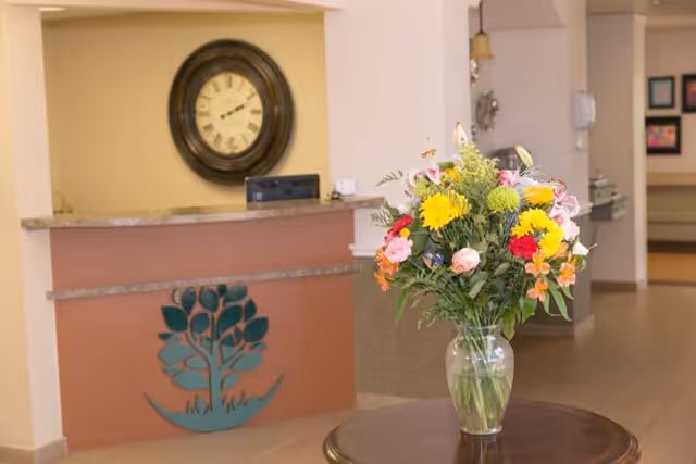 A reception area with a wooden desk featuring a decorative tree design on the front. Behind the desk is a large round wall clock. In the foreground, there is a round wooden table with a clear glass vase holding a colorful bouquet of flowers including yellow, pink, red, and white blooms. The background shows a hallway with framed pictures on the wall.