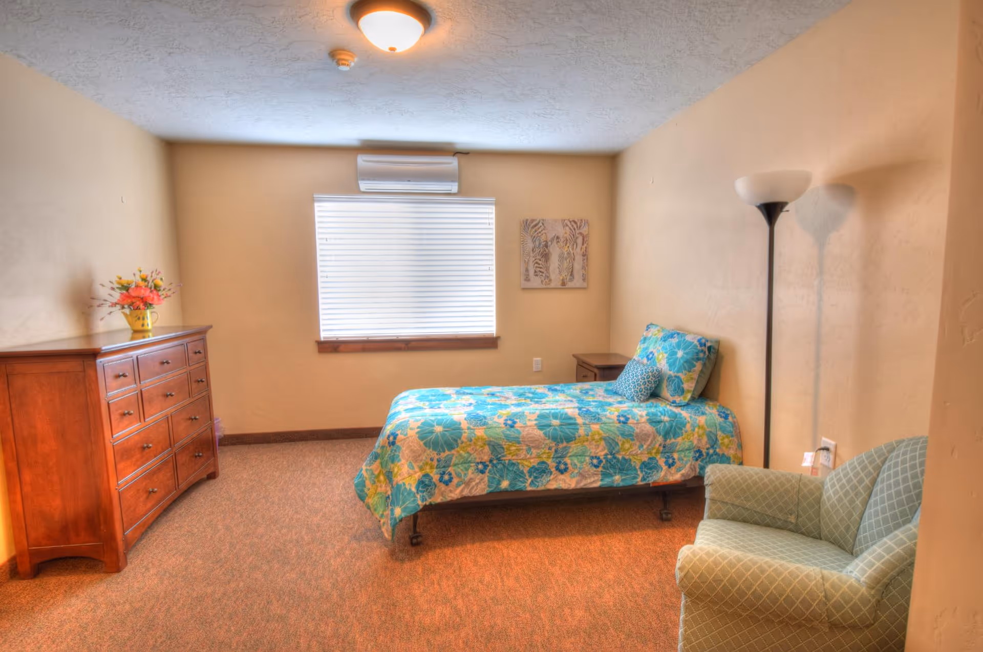 A bedroom with a single bed covered in a blue and green floral quilt, a wooden dresser with a vase of flowers on top, a small wooden nightstand, a green upholstered armchair, a floor lamp, and a window with closed blinds and an air conditioning unit above it.