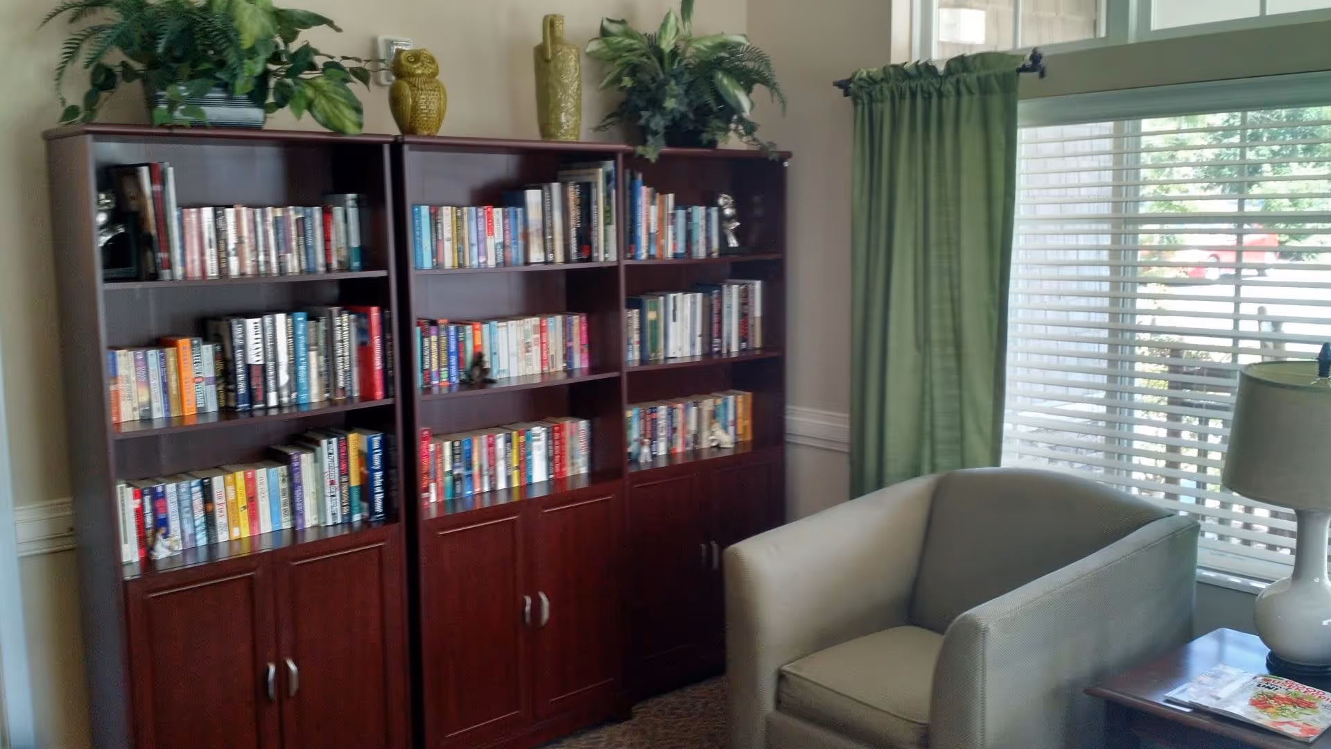 A cozy reading corner in a senior living facility featuring two wooden bookshelves filled with books and decorative items on top, a comfortable armchair, a side table with a lamp, and a window with green curtains and blinds.