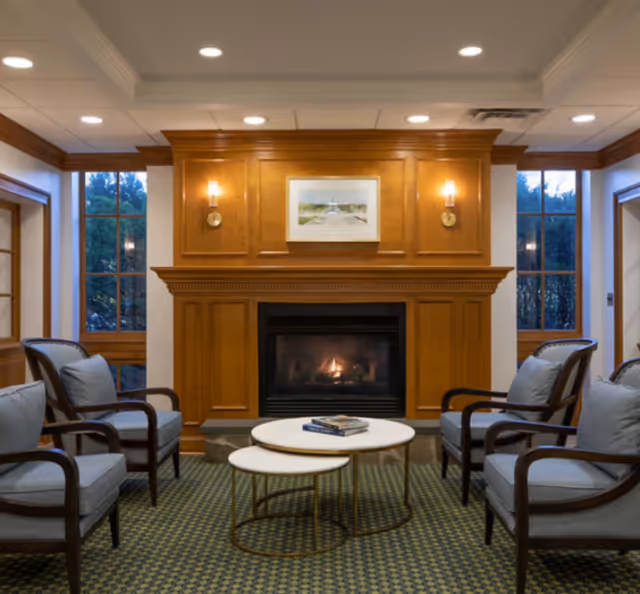 A cozy sitting area with four cushioned armchairs arranged around two nested round coffee tables in front of a wooden fireplace with a fire burning. The fireplace is flanked by two wall sconces and a framed picture above it. Large windows on either side let in natural light.