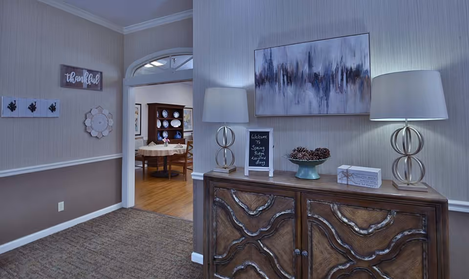 Interior view of a hallway in Spring Ridge Assisted Living featuring a wooden sideboard with two modern lamps, a decorative bowl with pinecones, a small chalkboard sign that reads 'Welcome to Spring Ridge Assisted Living,' and a piece of abstract wall art above. The hallway leads into a dining area with a round table and chairs, and a wooden cabinet displaying plates. The walls are decorated with framed art and a sign that says 'thankful.'
