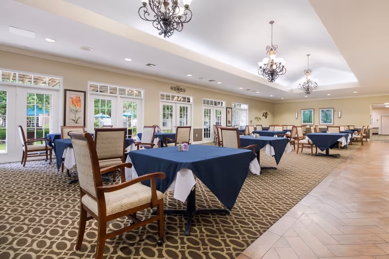 Spacious dining room with multiple tables covered in dark blue tablecloths, wooden chairs, chandeliers, and large windows overlooking greenery.
