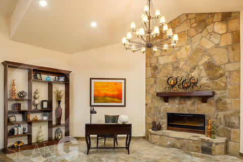 A cozy interior room featuring a stone fireplace with decorative items on the mantel, a wooden desk with a chair, a framed painting on the wall, and a tall wooden bookshelf with various decorative objects and books.