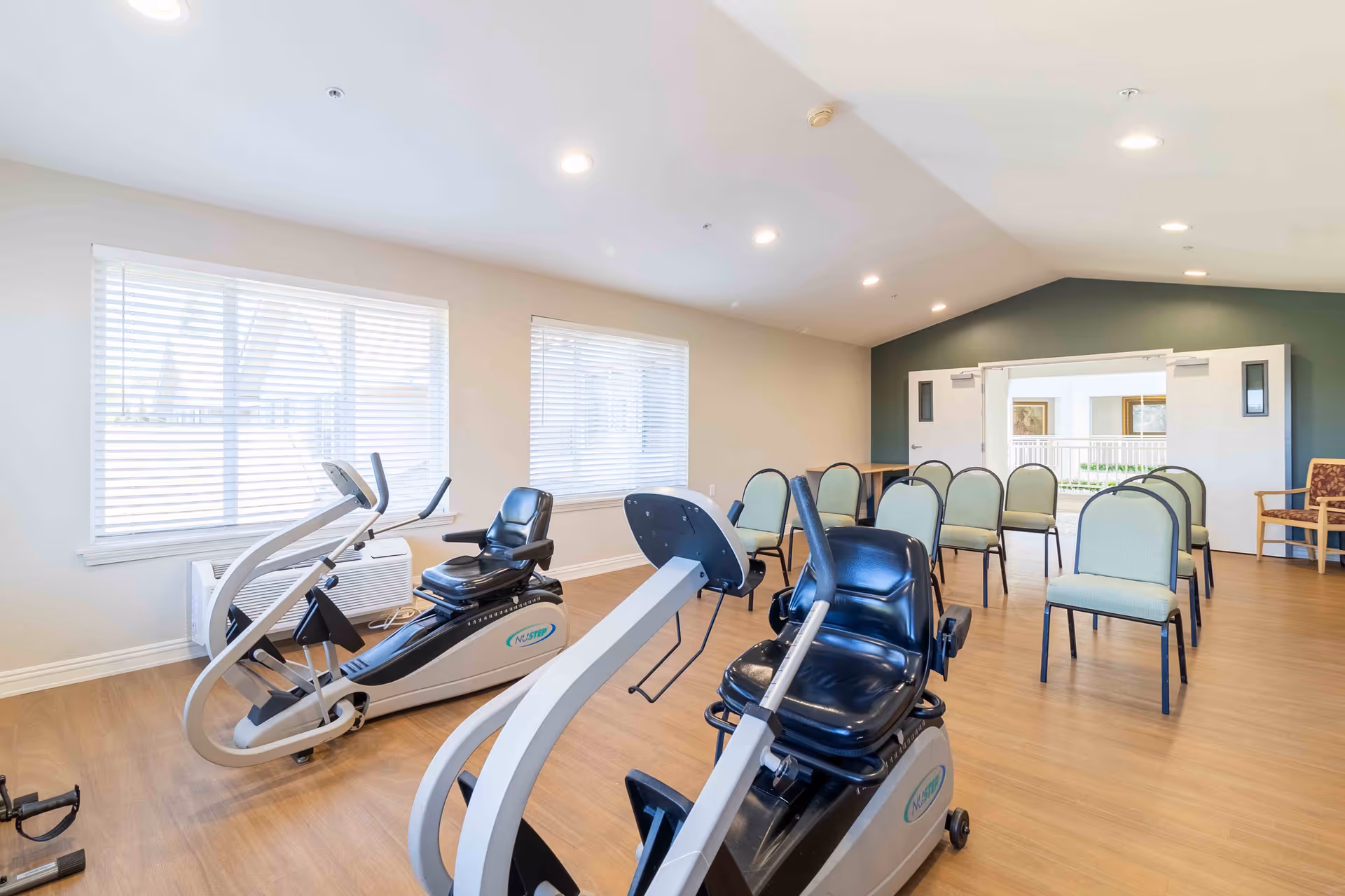 A bright room with exercise equipment including two recumbent stationary bikes in the foreground and several chairs arranged in rows in the background. The room has large windows with blinds, light-colored walls, and wood flooring.