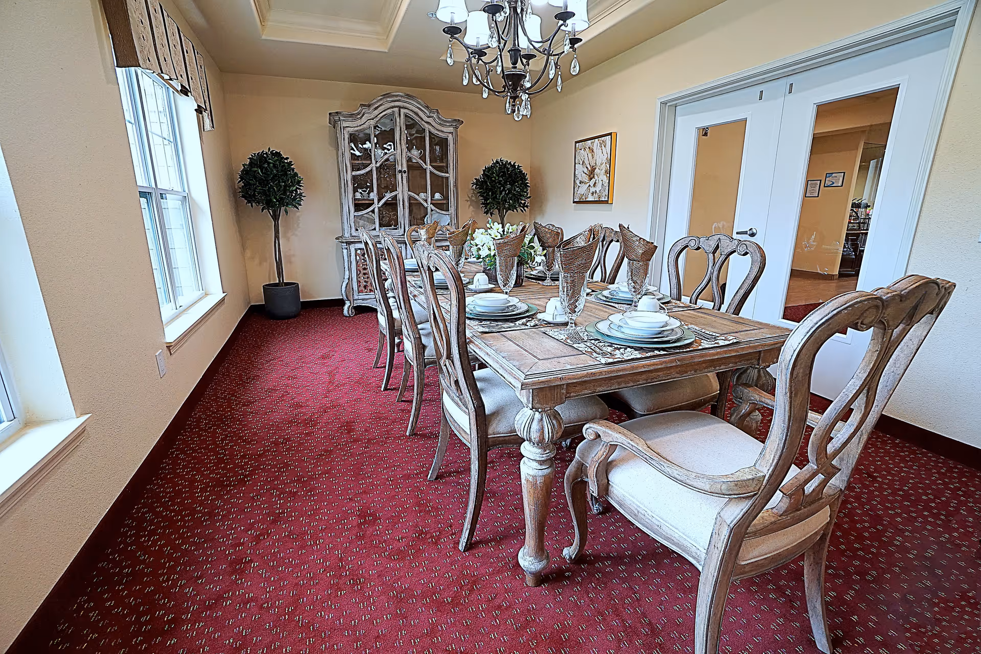 A formal dining room with a long wooden dining table set for eight people with plates, glasses, and napkins. The room has red carpet flooring, two large windows on the left, a chandelier hanging from the ceiling, two potted plants in the back corners, a decorative cabinet against the far wall, and a framed artwork on the right wall.