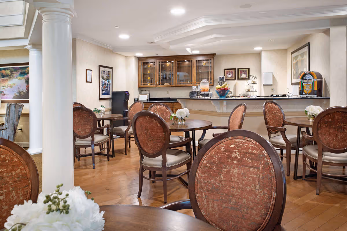 A cozy dining area with round wooden tables and cushioned chairs arranged neatly. The room features hardwood flooring, white columns, and soft lighting from ceiling fixtures. In the background, there is a counter with a glass cabinet above it, displaying various items, and a small jukebox on the right side. The walls are decorated with framed pictures and artwork.