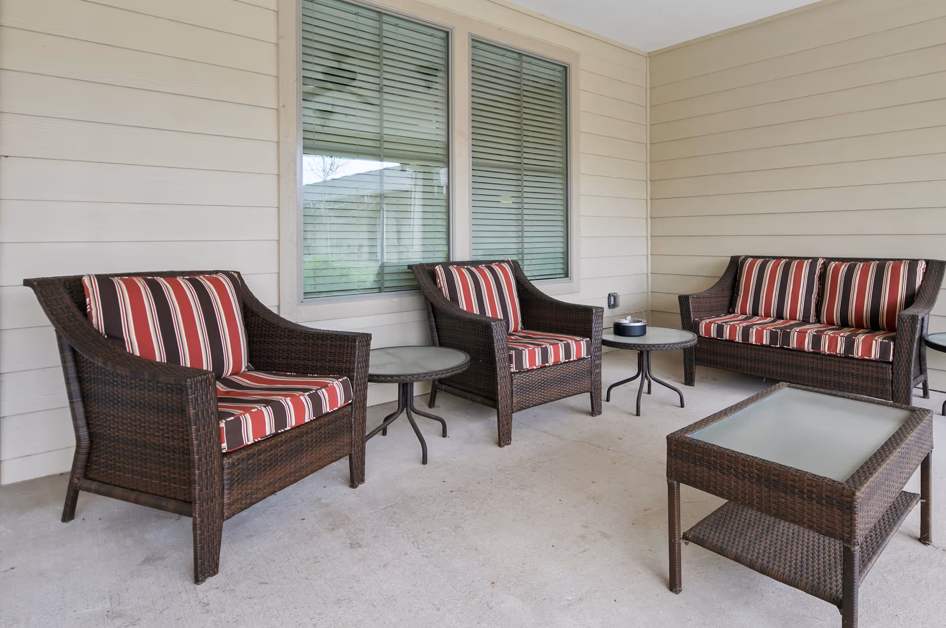 Outdoor covered patio area with brown wicker furniture including two armchairs, a loveseat, and a coffee table, all with red, white, and brown striped cushions. There are also two small round side tables with glass tops. The patio has beige siding walls and a window with closed blinds.