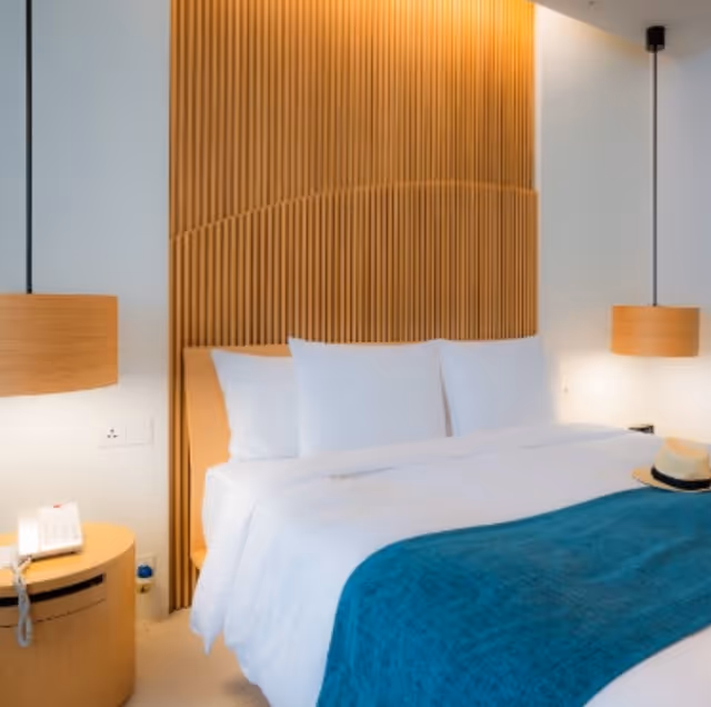 A neatly made bed with white pillows and sheets, topped with a teal blanket. The bed has a wooden headboard with vertical slats and warm lighting above it. On each side of the bed, there are hanging wooden pendant lamps. A small round wooden bedside table with a telephone and a hat placed on the bed are also visible.
