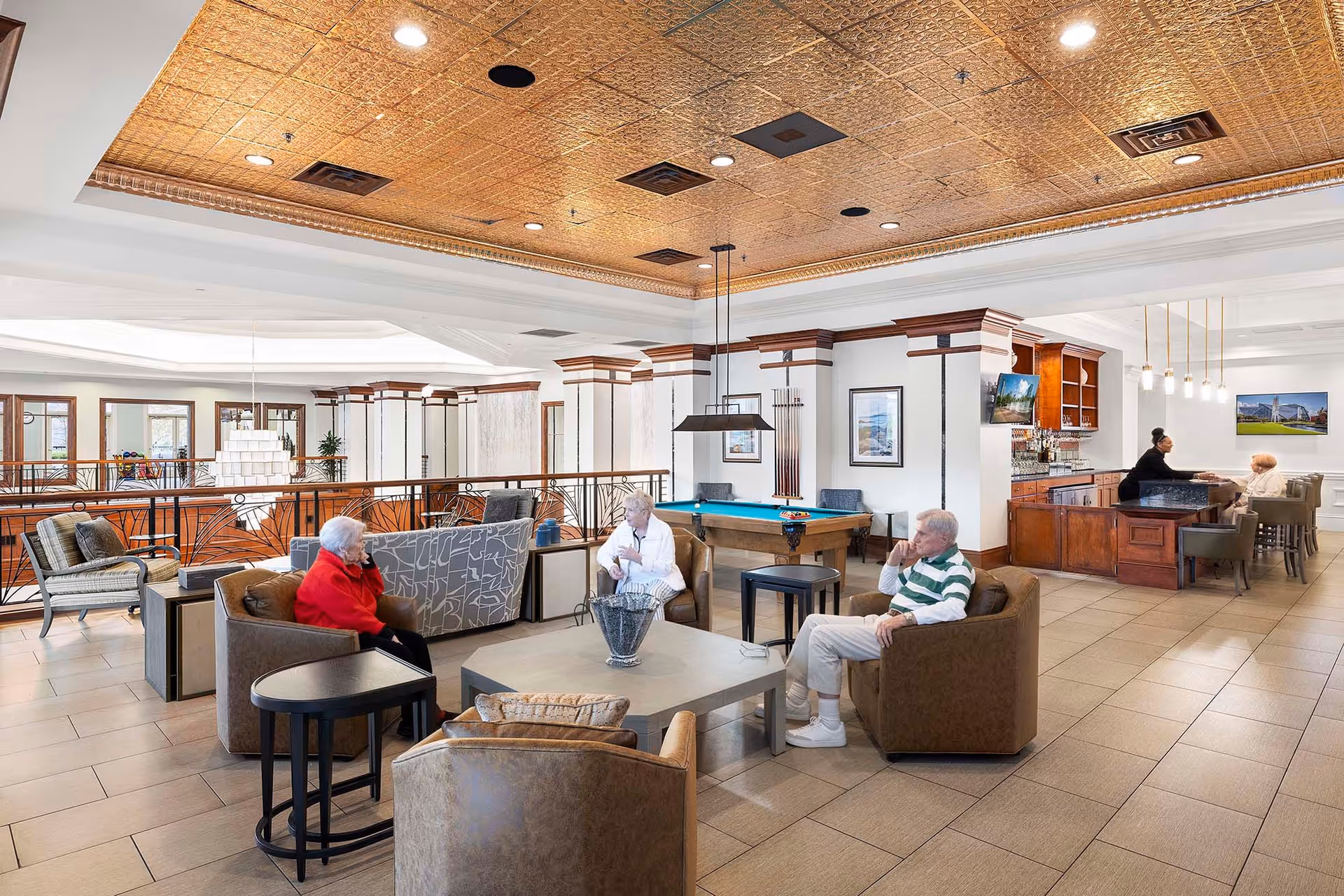 Bright communal lounge in a senior living facility with sofas and chairs arranged around a coffee table, a pool table, and a bar area.