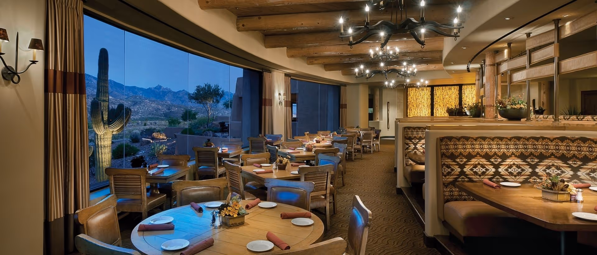 Cozy Southwestern-style dining room with round tables and booths, wooden chairs, chandeliers, and large windows overlooking a desert landscape with a saguaro cactus.