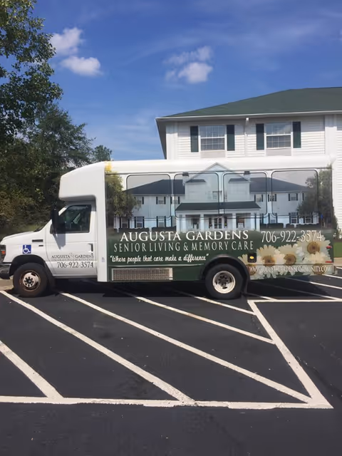 A white Augusta Gardens senior living shuttle van parked in a marked lot in front of a two-story building.