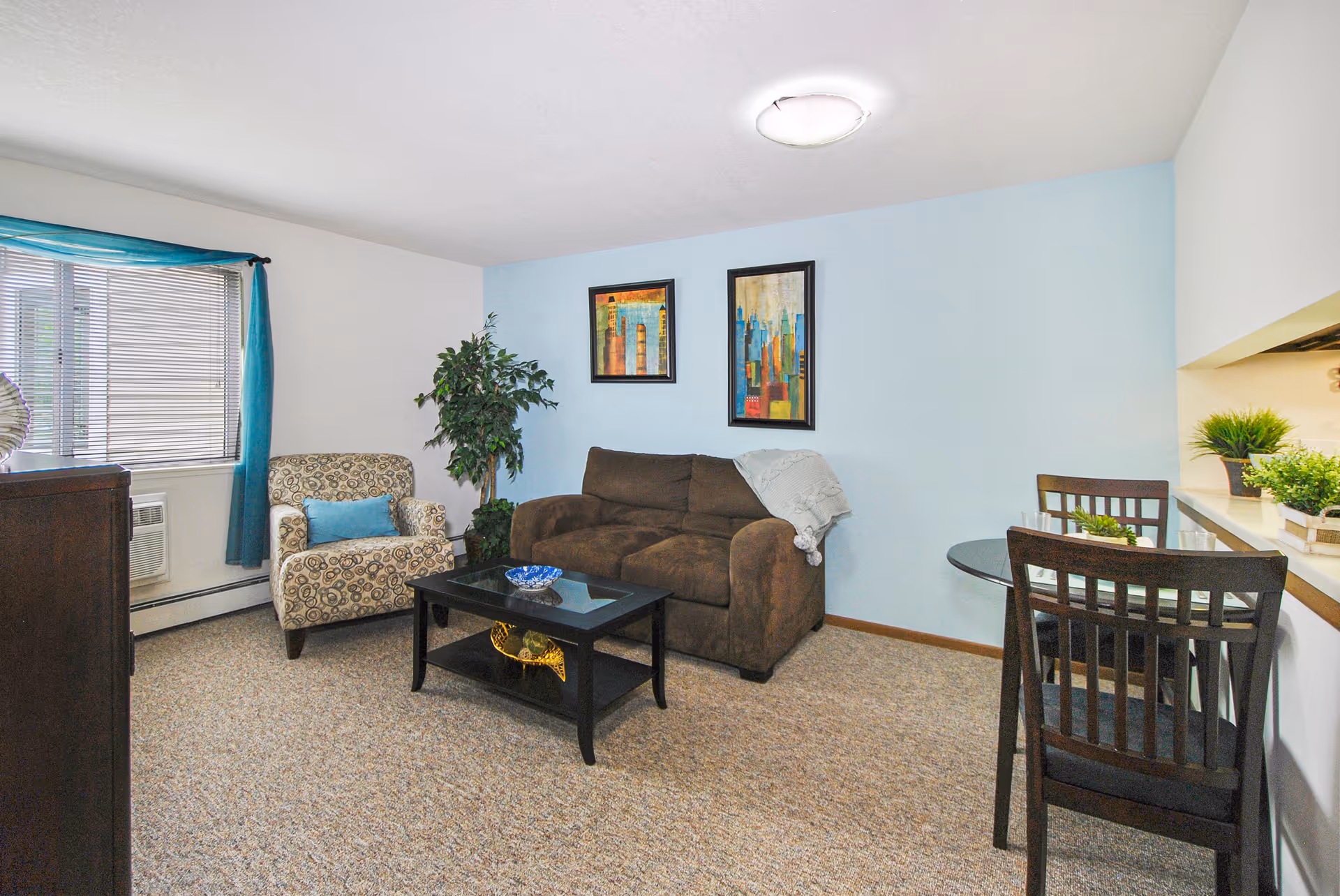 Well-lit living room with a brown sofa, patterned armchair, coffee table, small dining table, and wall art.
