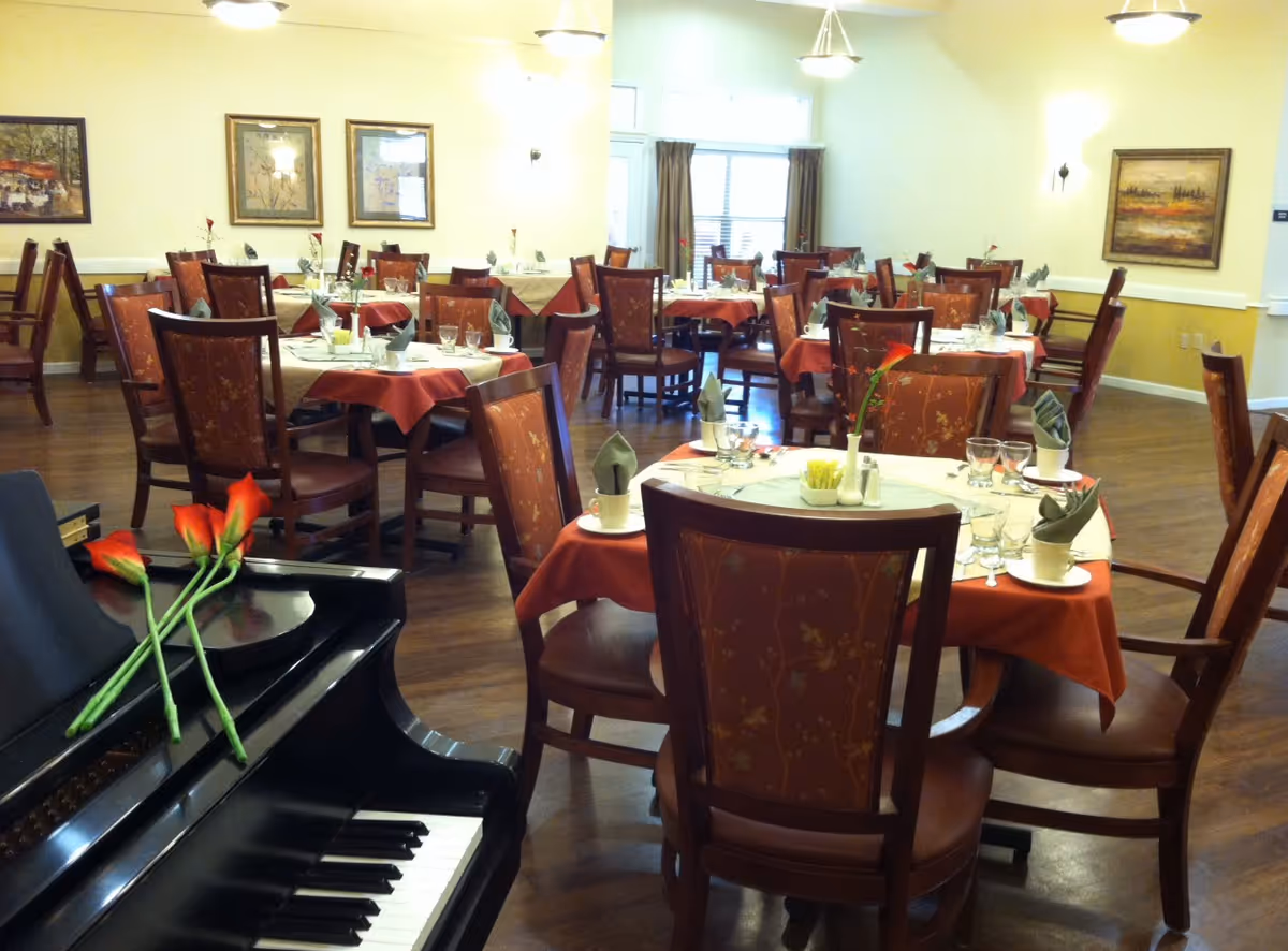 A dining room with multiple round tables covered with red and beige tablecloths, each set with glasses, cups, napkins, and a single flower in a vase. There are wooden chairs with patterned upholstery around the tables. In the foreground, there is a black piano with three red flowers placed on it. The room has wooden flooring, light yellow walls, framed artwork, and ceiling lights.