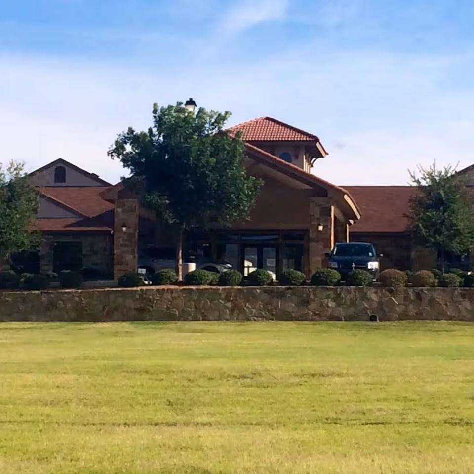 Front exterior view of Hilltop Park Rehabilitation And Care Center showing a stone and brick building with a red tiled roof, surrounded by trees and bushes, with a green lawn in the foreground under a blue sky.
