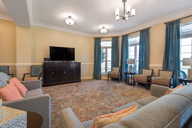 A spacious living room with beige walls and carpeted floor featuring a large area rug. The room has multiple seating options including sofas with orange and beige pillows, armchairs, and side tables with lamps. A flat-screen TV is placed on a dark wooden cabinet against one wall. Large windows with blue curtains allow natural light to fill the room.