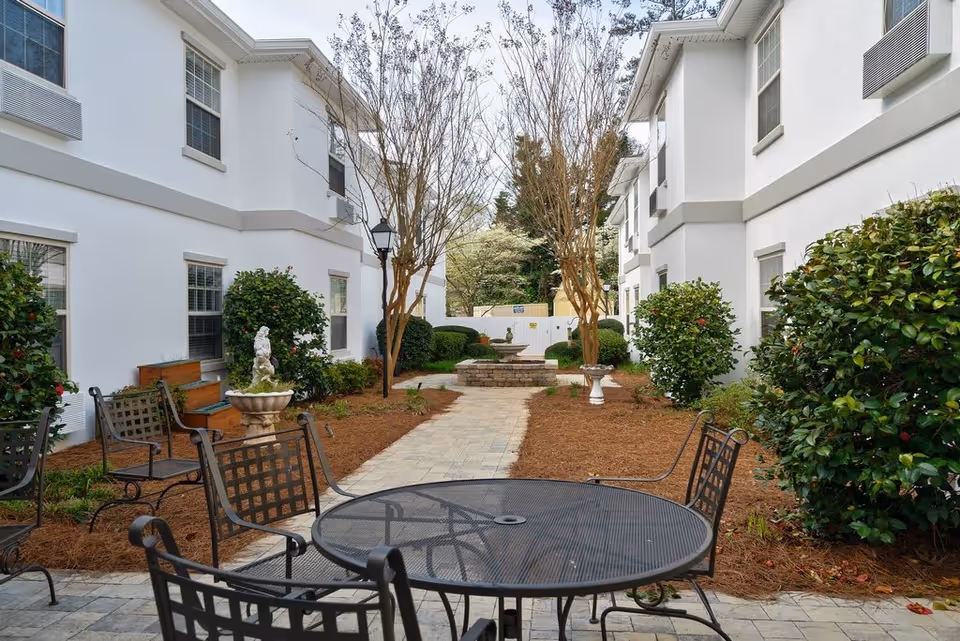 Outdoor courtyard area between two white buildings with a paved walkway, metal patio table and chairs, decorative fountains, bushes, and trees.