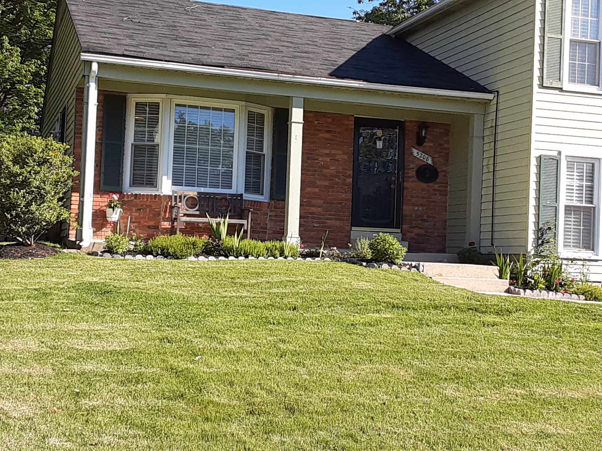 Front exterior view of a single-story brick and siding house with a small porch, a bench, green shutters, and a well-maintained lawn with shrubs and plants.