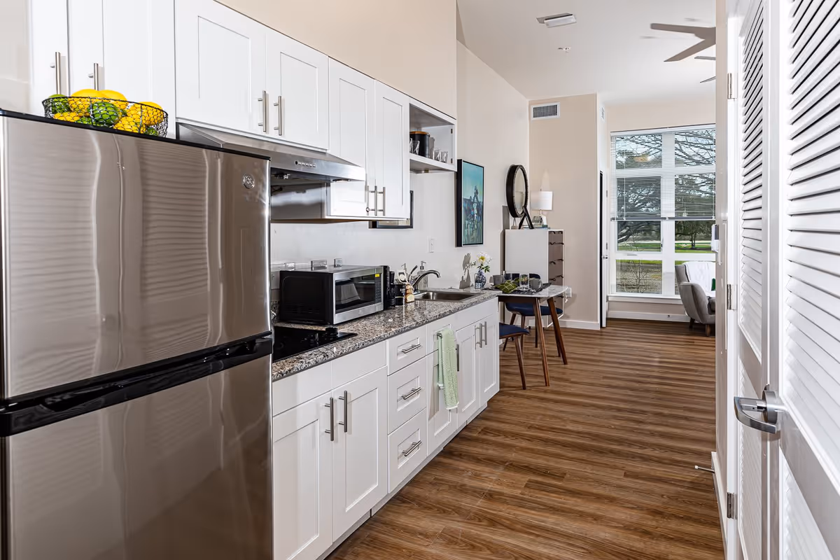 A modern kitchen area with white cabinets, a stainless steel refrigerator, microwave, and a granite countertop. The kitchen opens into a small dining area with a table set for two, a dresser with a round mirror, and a large window letting in natural light. The floor is wood with a warm tone.