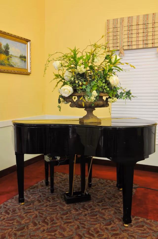 A black grand piano topped with a large floral arrangement sits on a patterned rug in a yellow-walled room.