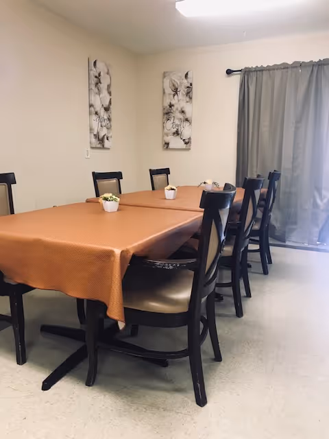 A dining room with two rectangular tables pushed together, covered with a brown tablecloth. There are eight dark wooden chairs with beige cushions around the tables. Small potted plants are placed on the tables. The room has light-colored walls with two floral paintings and a window covered by gray curtains.