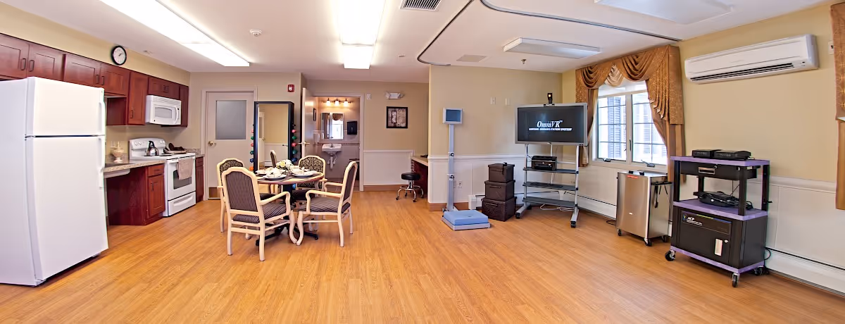 A spacious room with a kitchen area on the left featuring a white refrigerator, stove, microwave, and wooden cabinets. In the center, there is a round dining table set with four cushioned chairs. The right side of the room has a television on a stand, a window with brown curtains, and various electronic equipment. The room has wood flooring, beige walls, and ceiling lights.