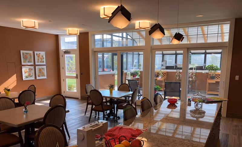 Communal dining area with several tables and chairs, a long countertop island in the foreground, and large windows/doors leading to an enclosed patio.