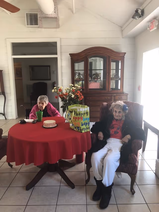 Two elderly women sitting in a living room area of an assisted living facility. One woman is seated at a round table covered with a red tablecloth, with a small cake, a green cup, and a gift bag on the table. The other woman is sitting in an armchair next to the table. Behind them is a wooden cabinet with glass doors displaying decorative items. The room has tiled floors and white walls with a door leading to another room.