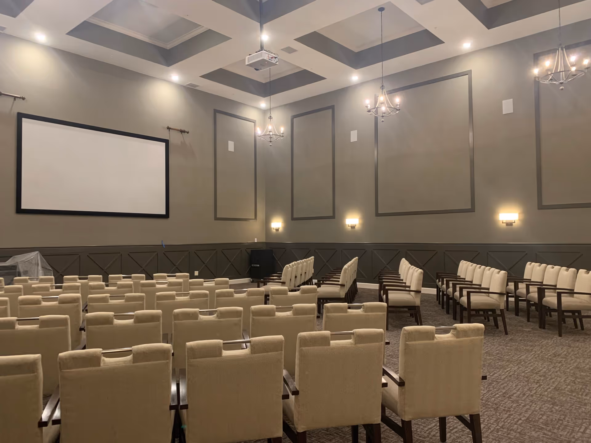 Large multipurpose auditorium with rows of beige upholstered chairs facing a blank projection screen on a gray paneled wall.