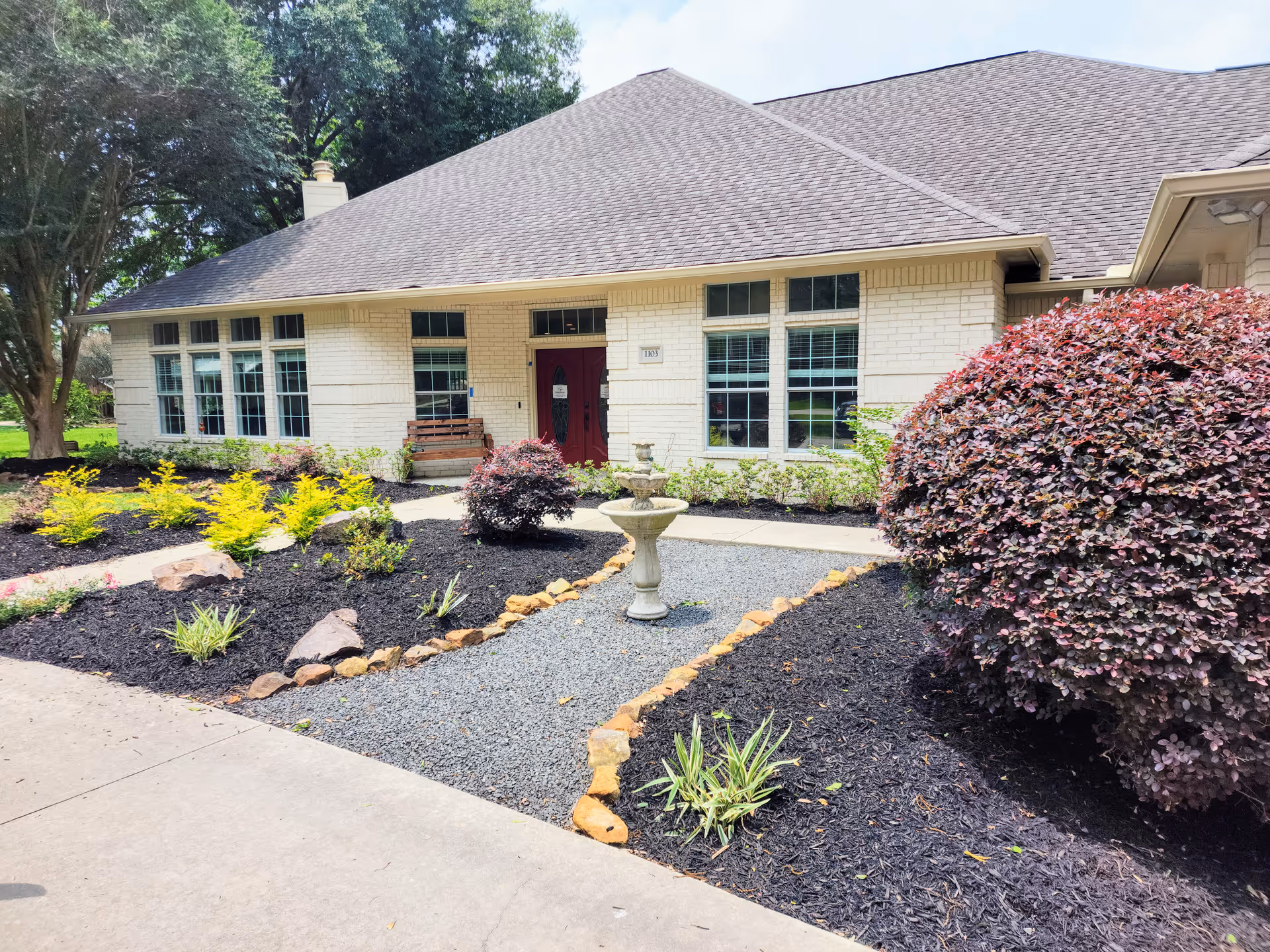 Exterior view of a single-story brick building with a large roof, multiple windows, and a red double door entrance. The front yard features a landscaped garden with various shrubs, a small stone-lined gravel path, and a decorative birdbath or fountain in the center. Trees and greenery surround the building under a partly cloudy sky.