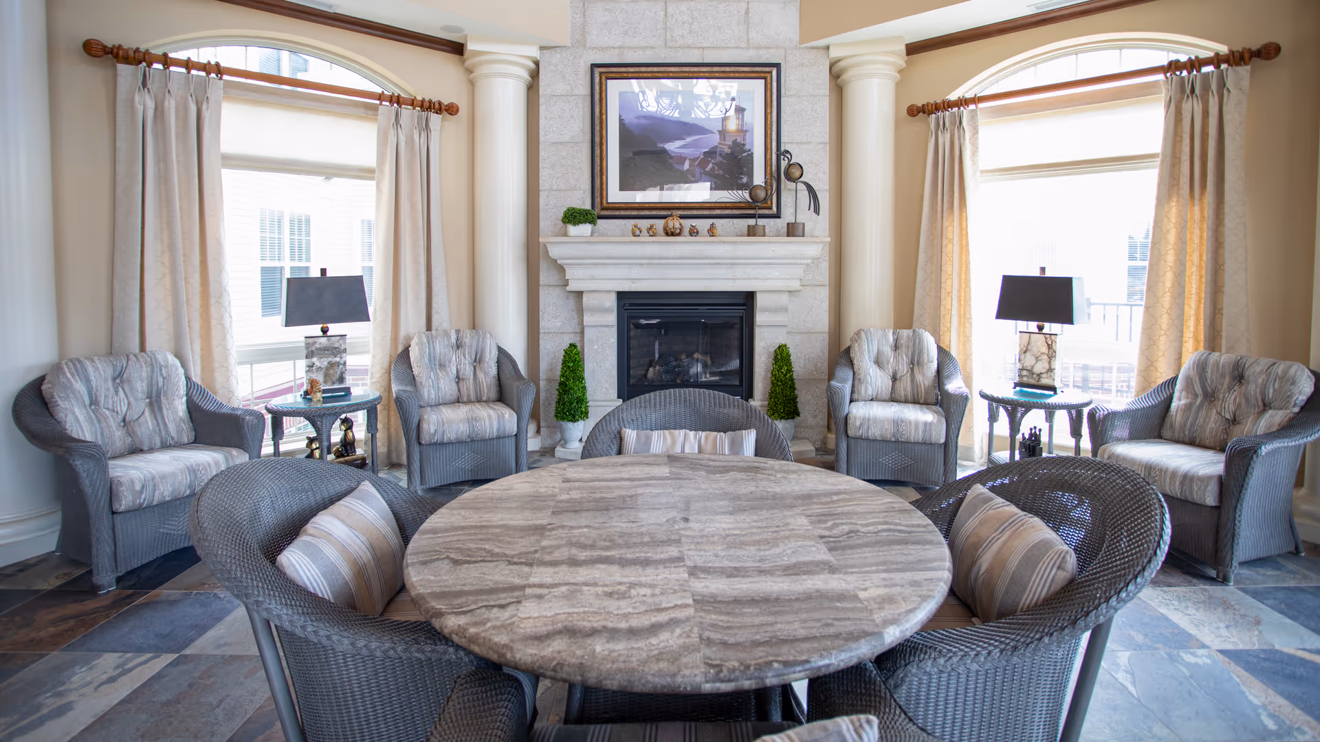 Bright senior living sitting area with a round stone table, wicker armchairs, and a fireplace between two tall windows.