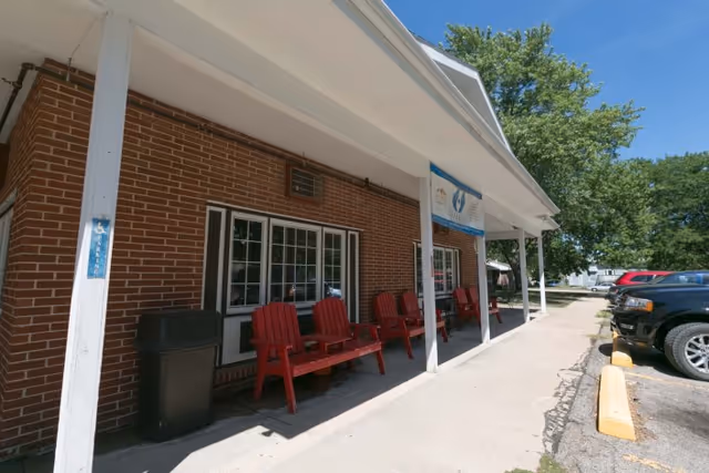 Exterior view of Beatrice Health and Rehabilitation facility showing a brick building with a covered porch. The porch has several red wooden chairs and a bench. There is a trash can near a white column with a handicap sign. Several cars are parked along the driveway beside the building, and trees are visible in the background under a clear blue sky.