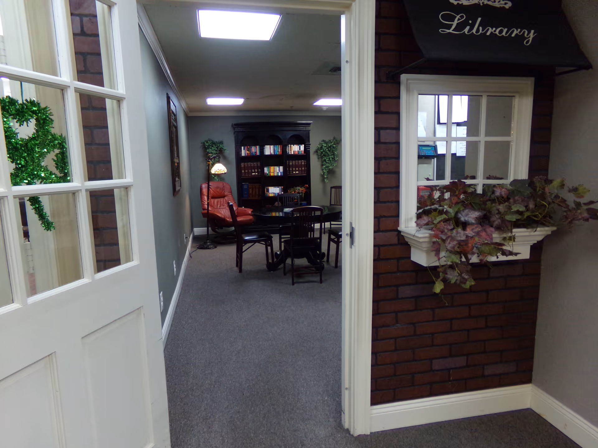Interior view through a doorway into a small library/lounge area with bookshelves, a table and chairs, and a red armchair.