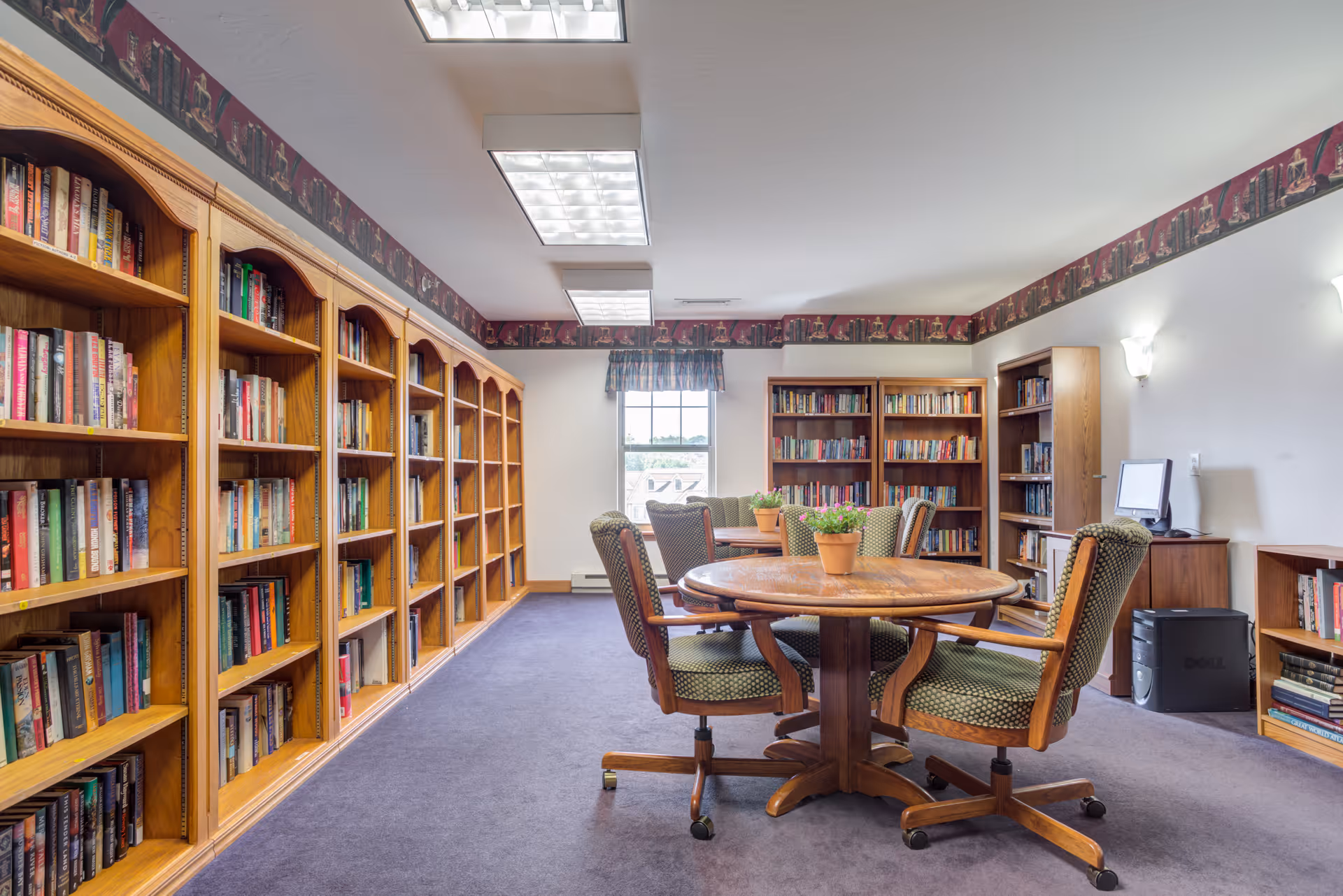 A cozy library room with wooden bookshelves filled with books lining the left and back walls. In the center, there is a round wooden table surrounded by four green upholstered chairs on casters. A window with a valance lets in natural light, and a computer workstation is visible on the right side of the room. The room has a purple carpet and fluorescent ceiling lights.