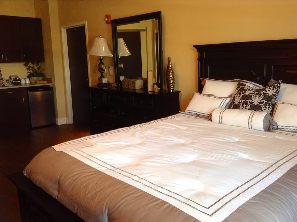 A neatly made bed with white and beige bedding and multiple pillows in a senior living facility bedroom. Next to the bed is a dark wooden dresser with a large mirror, a decorative lamp, and some decorative items. In the background, there is a small kitchenette area with dark cabinets and a mini refrigerator.